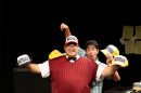 A man in a red sweater vest, a black bowtie, and khakis wears a red trucker hat that says "Monk" and holds two other trucker hats. Behind him, a man in a short sleeved plaid button up holds another trucker hat in one hand and a remote in the other. 
