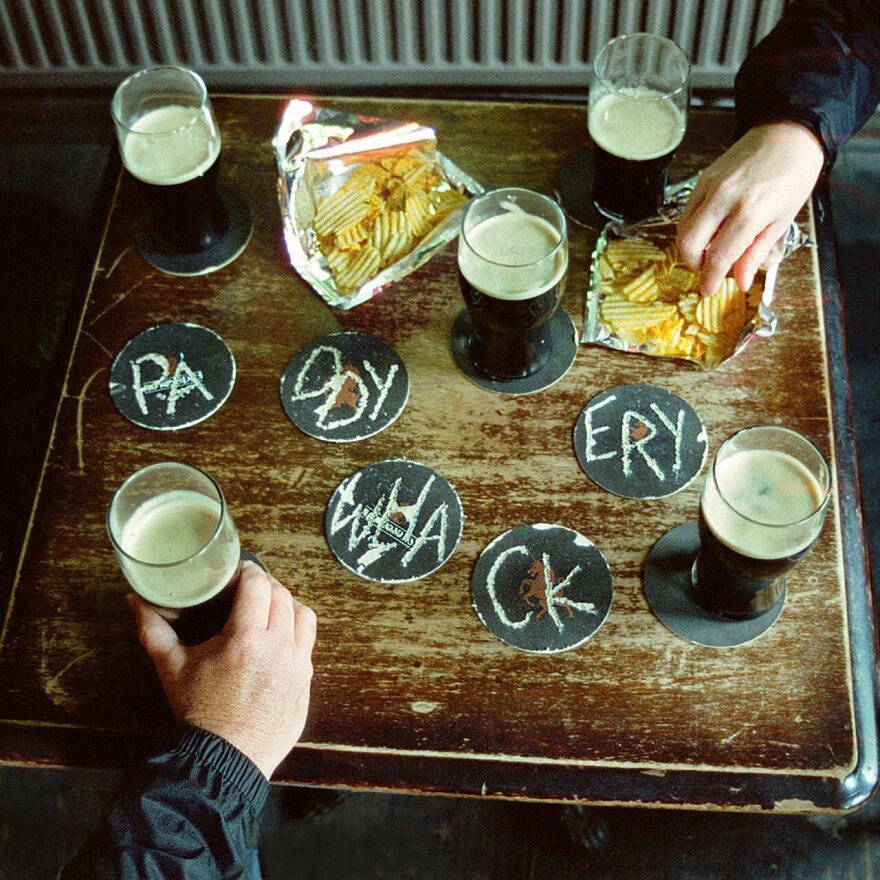 A worn wooden tabletop with coasters, pints of beer and chips. There are two hands that are only visible from the wrist area towards the fingers, hlding beer glasses. On coasters with no beer on them the word "PA DDY WHA CK ERY" or "paddywackery" is spelled out across five coasters.