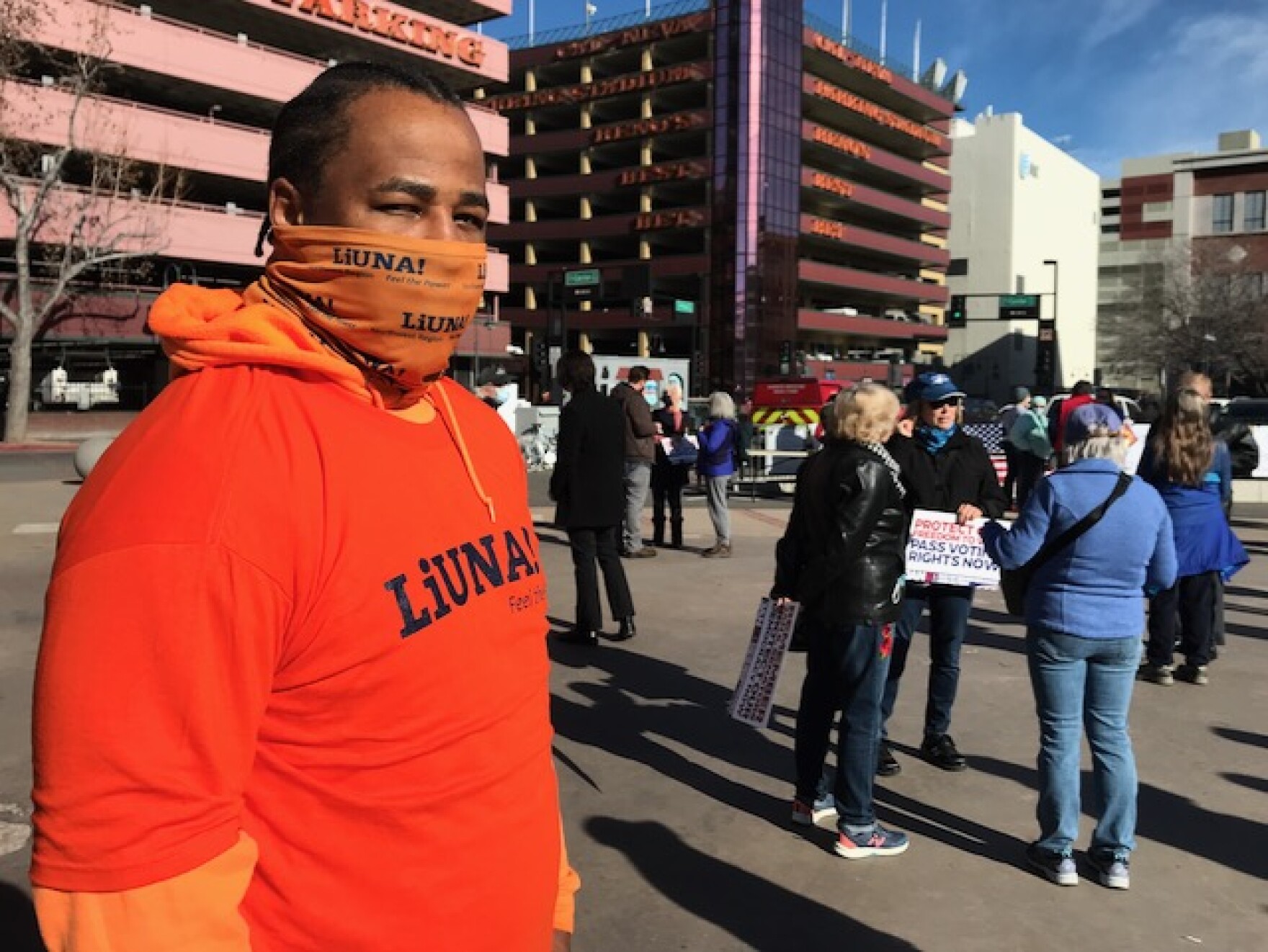 King Dupree wears an orange shirt and face mask. He stands at the left side of the frame. Other people fill the background.