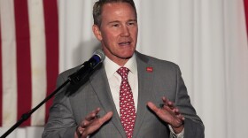 a man in a gray suit and red tie speaks in front of a microphone while gesticulating with his hands