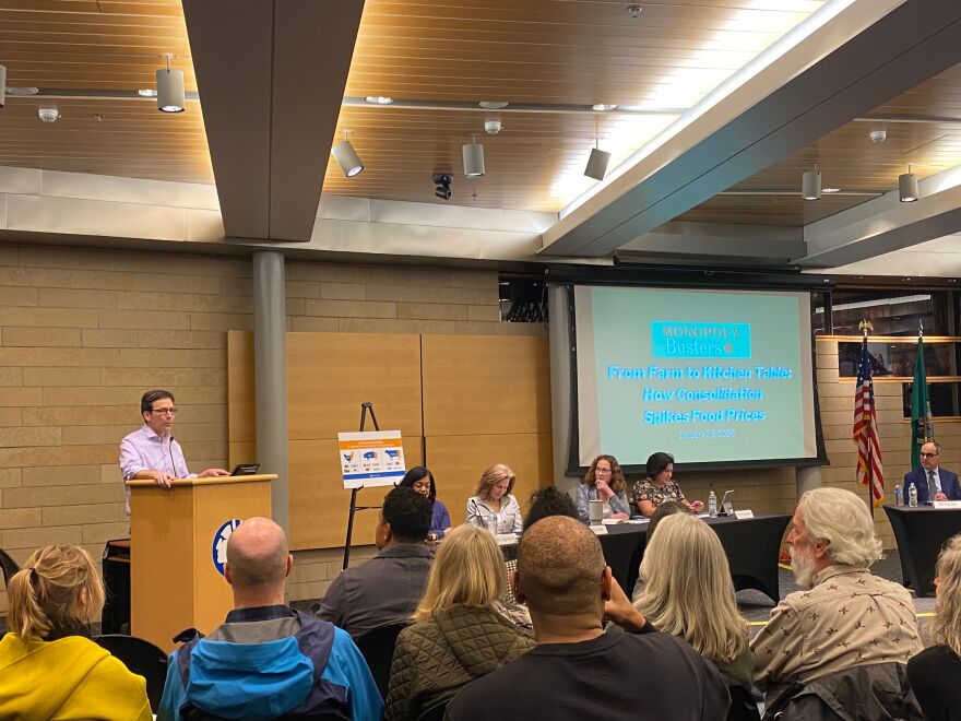 Washington Governor Bob Ferguson speaks to a crowd from behind a lectern. He is next to a table where several lawmakers are seated. A screen in the back displays a presentation titled "From farm to kitchen tables: How consolidation spikes food prices." 