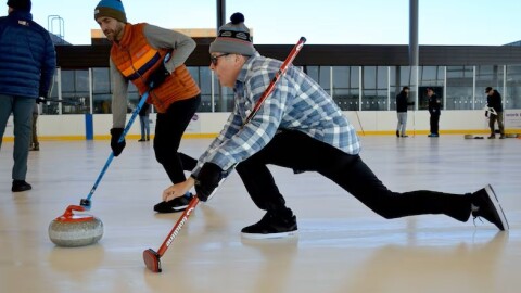 Two men play curling on an outdoor ice rink, with one sliding forward in a low lunge while releasing a granite stone and the other sweeping the ice ahead of it with a broom.