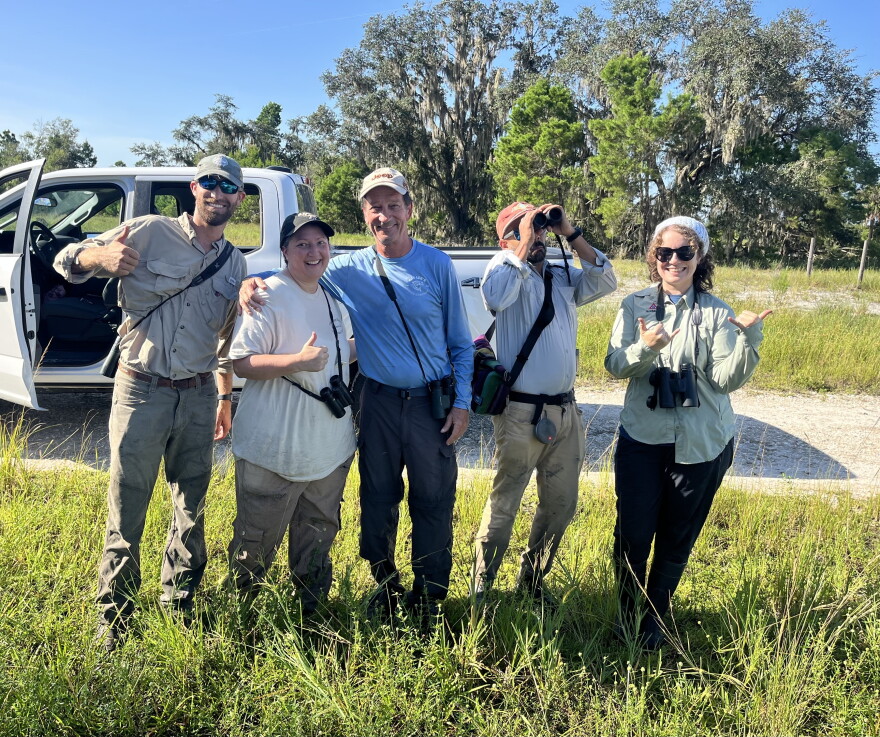 5 people give a thumbs up standing outdoors, smiling.