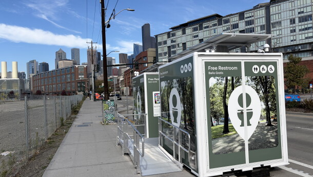 A photo of two Throne bathrooms with the Seattle skyline visible in the background. The bathroom trailers have images of trees on the outside and signage that says "free restroom" and an image of a toilet. 
