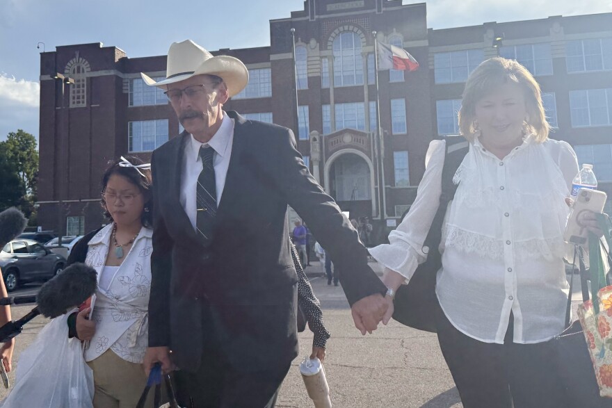 A man with a mustache wearing a suit, glasses and a cowboy hat walks holding hands with a woman in a white blouse. 