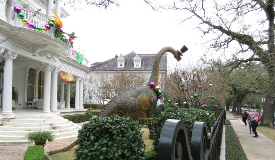 A small group of people stand on a sidewalk looking at Mardi Gras decorations in the front yard of a house. Behind the yard's wrought-iron fence stands a giant Apatosaurus-like dinosaur wearing a top hat. Purple, green and gold balls hang from a leafless tree in the yard, and purple, green and gold oversized garland hangs on the white colonial-style balustrade on the house.