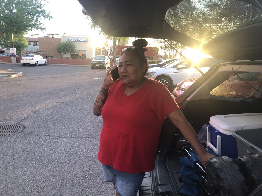 A woman speaks on a cell phone in front of an open car trunk with a cooler in