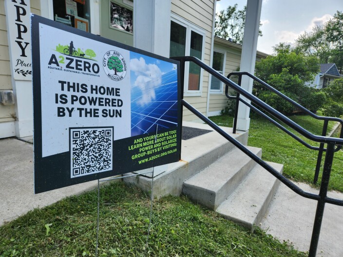 A photo of a sign in front of a building. The sign says "A2Zero" and "This home is powered by the sun and yours can be too! Learn more about solar power and Solarize group-buys by visiting www.a2gov.org/solar."