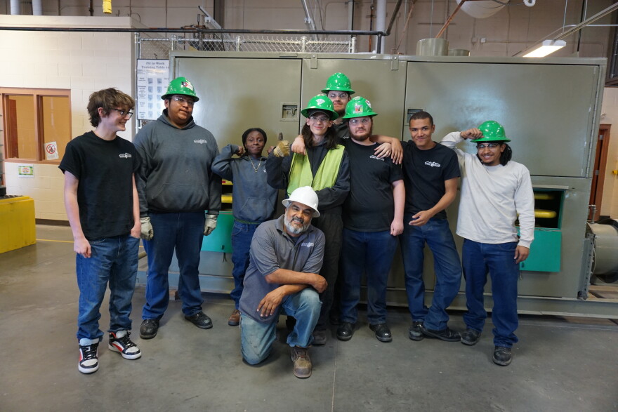 Eight young adults, some in green hardhats, pose around an older man in a space that looks like a warehouse.