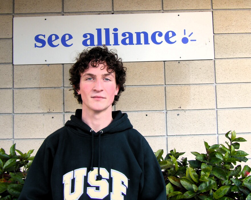 Man in a USF hoodie standing in front of a sign that says "see alliance"