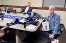 Two smiling men sitting at a table in a radio talk studio: the man back left has short dark hair and a dark mustache and is wearing a white button-down shirt under a burgundy suit; a man front right has very short dark hair and is wearing a blue button-down shirt, jeans, blue socks, and white and red sneakers. He is holding a book titled "The Age of Extraction." 