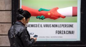 A woman, wearing a respiratory mask, walks past a placard showing a handshake between two hands representing the Chinese and the Italian national flags and reading 'The enemy is the virus, not the people. Go China!' in the Paolo Sarpi district (Milan's Chinatown) in Milan, Italy.