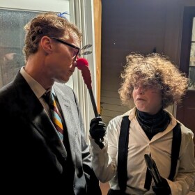 A tall man with a red clown nose looking down at a woman who is smirking and grabbing his nose with some tongs