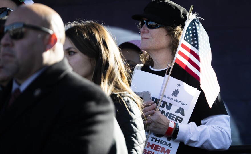 A woman holds a U.S. flag and sign as she stands in a crowd. 