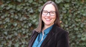 a white woman with dark hair and glasses wearing a teal colored shirt and brown velvet coat smiles and stands in front of a dark ivy background