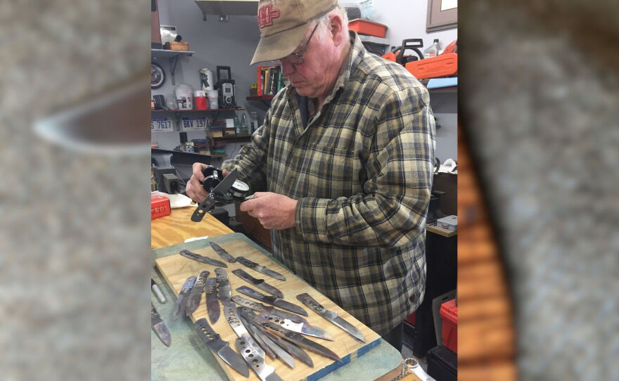 A man wearing a plaid patter shirt measuring silver knives with a table with a pile of knives in front of him.