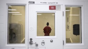 A person in a prison jumpsuit stands behind a door with a glass window.