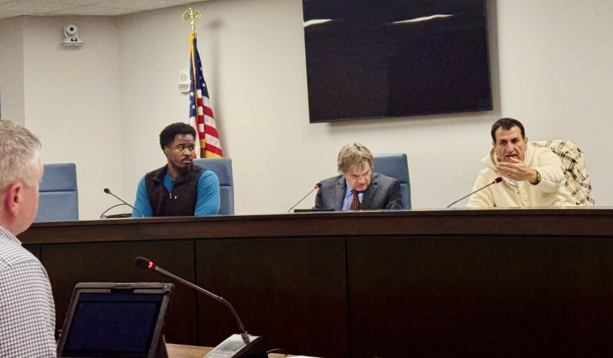 Three men sit at a government meeting table. One man speaks and gestures with his hand, another looks forward, and the third reads documents. An American flag and a large monitor are on the wall behind them.