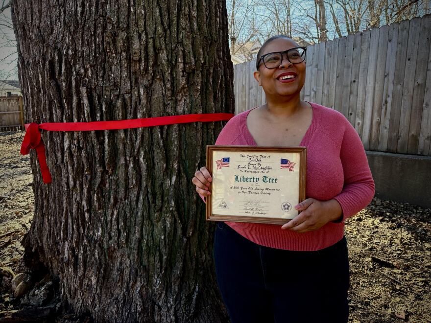 A woman in a pink shirt holds a certificate in front of a large tree.