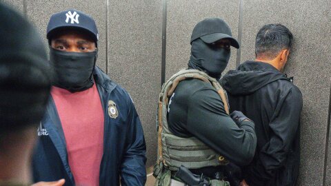 A man wearing handcuffs is pinned to the wall by a masked man wearing a tactical vest and a gun while another stands nearby.