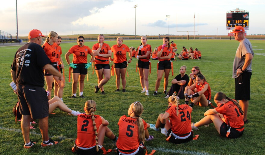 Two men standing at either side of a circle of high school girls -- some standing and some sitting -- on a high school field. 