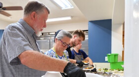 four servers prepare meals in a kitchen. the man closest to the camera with a white beard and hair in a gray short sleeve collared shirt has a stack of plastic plates and bowls. the others behind him are filling the plates and bowls with food