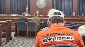 A person in an orange shirt reading "our democracy is not for sale" sits facing away from the camera as a meeting proceeds at the front of a carpeted room.