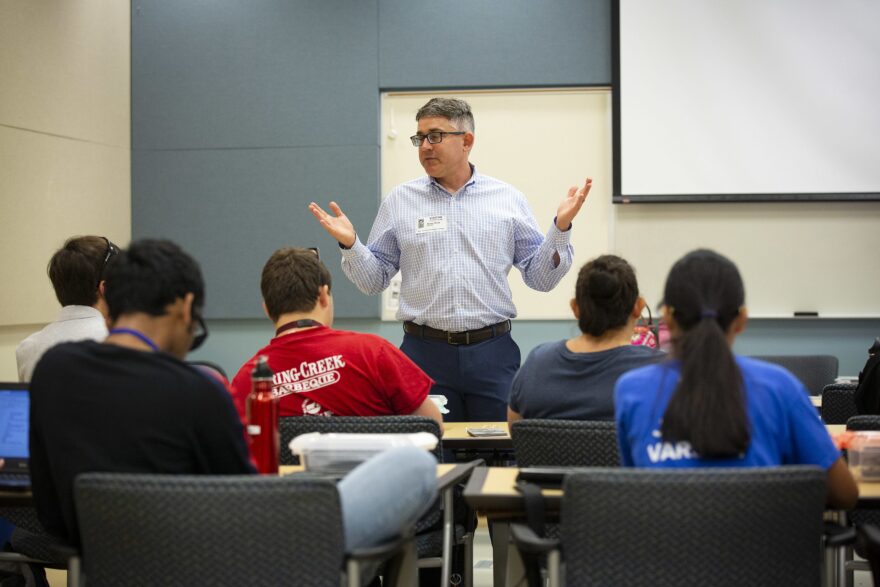 A man stands in front of a group of students. The students are seen from behind and the man is facing them, arms raised while speaking.