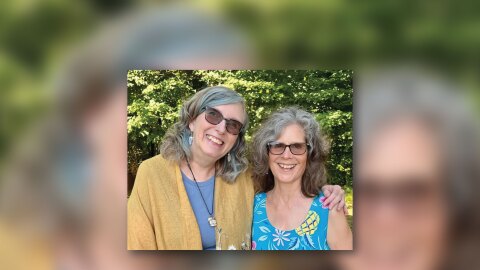 Two women pose next to each other. They wear brightly colored floral shirts. 