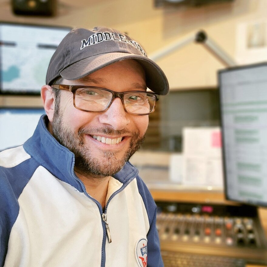 Mitch Teich, pictured in a quarter-zip, tortoise shell glasses and a baseball cap smiles in front of radio equipment. 