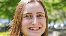 Emyle is pictured smiling at the camera. They are a white person with long brown hair and brown eyes. They are wearing a black blouse and standing outside. 