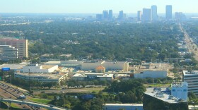 an aerial image showing a large interior shopping mall in the foregrown and downtown skyline through hazy sky toward the rear of the frame.  A major road on the right of the mall and image frame 