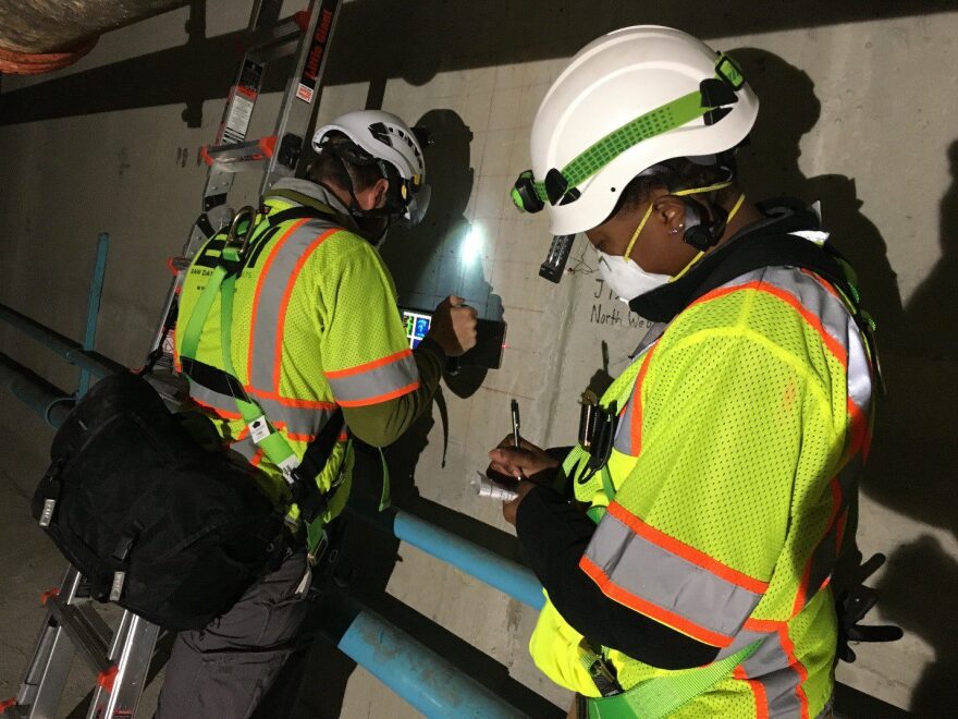 Two people in reflective gear and hard hats take notes and examine a concrete wall.