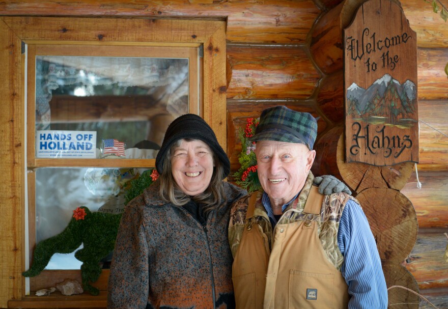 Shirley and LLoyd Hahn stand in front of their log cabin home, Dec. 13, 2022. A sticker on the window says "Hands off Holland." A carved wooden board hanging on the house says "Welcome to the Hahns". Shirley and Lloyd live in Condon, MT and operated a business constructing log homes for decades. Shirley says the couple has seen wildlife of all kinds pass through their backyard over the years. She fears that would end with a tripling of the traffic at an expanded Holland Lake Lodge.