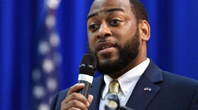 a black man in a suit and tie with a flag pin on his label holds a microphone