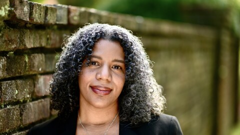 Shauna M. Morgan standing next to a weathered brick wall. She wears a black blazer and a necklace with a circle pendant. Her hair is long, curly, black and gray.