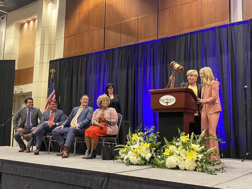 Four Fort Worth City Council members sit onstage, laughing as Mayor Betsy Price wields a huge novelty gavel at her podium. She has her arm around Mattie Parker, who is also laughing.