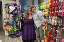 Two women lean into each other, smiling, in front of a wall with lots of colorful yarn hanging from racks