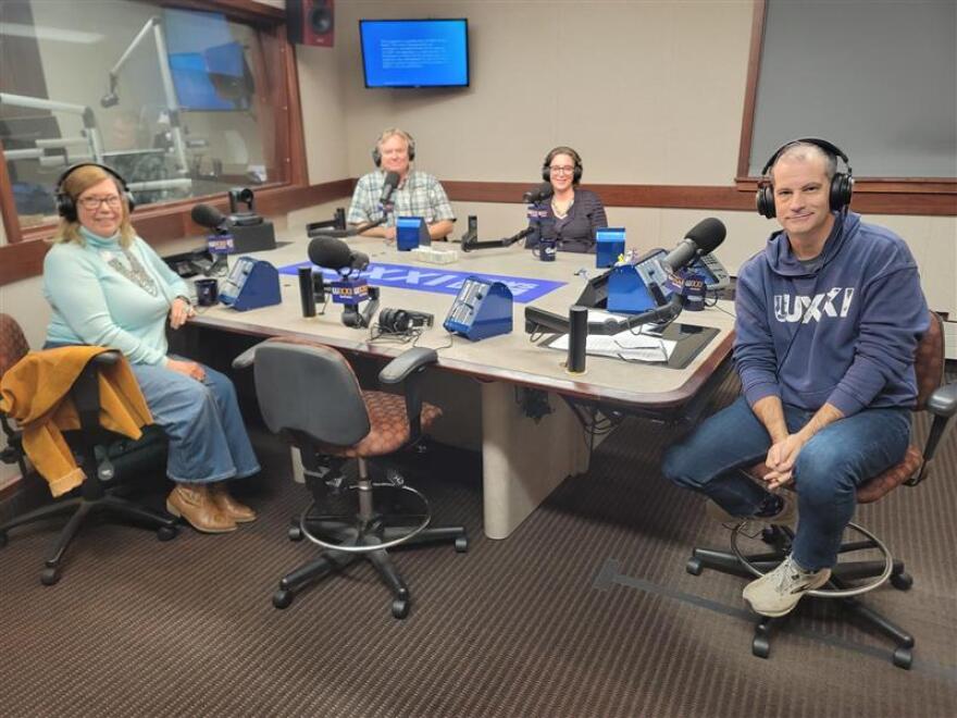 Four people wearing headphones sit at a table in a radio talk studio: a woman front left has long blonde hair and is wearing glasses, a light blue turtleneck sweater, a large multi-strand silver necklace, jeans and beige shoes; a man front right has short dark hair and is wearing a blue hooded sweatshirt with white lettering, jeans and sneakers; a man back left has short blonde hair and is wearing a short-sleeved plaid button-down shirt; a woman back right has short brown hair and is wearing glasses and a black blouse.