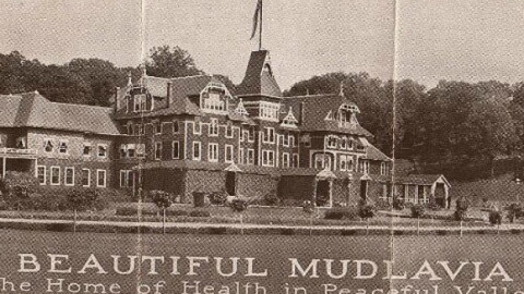 An archival publicity photo showing a large, multi-story Victorian-style hotel building with text at the bottom that reads 'BEAUTIFUL MUDLAVIA, The Home of Health in Peaceful Valley, KRAMER, INDIANA.'