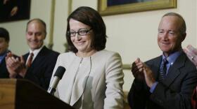 Loretta Rush stands smiling at a podium while Gov. Mitch Daniels, standing to her right, applauds during a formal ceremony.