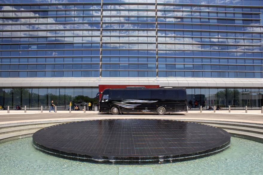A bus is parked in front of a windowed office building. The bus is faced to the left, and near the drivers side is a fountain that is in the shape of a circle that has lines cut through it, representing a wafer, the products TSMC makes that holds raw computer chips. 