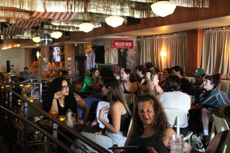 Customers at a bar sit around tables watching a soccer game.