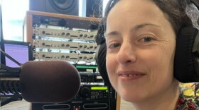 A woman wearing a floral blouse smiles in front of a microphone, various other pieces of radio equipment in the background