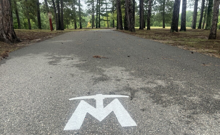 The Mesabi Trail logo is painted in white on the asphalt trail. Trees are visible in the background. 