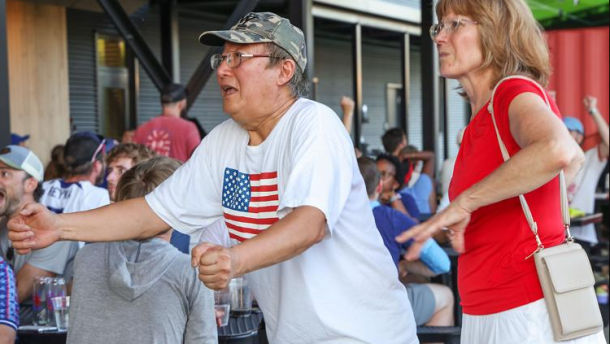 A man in a U.S. flag tee shirt and a woman in a red shirt and white skirt watch a soccer match at a bar.