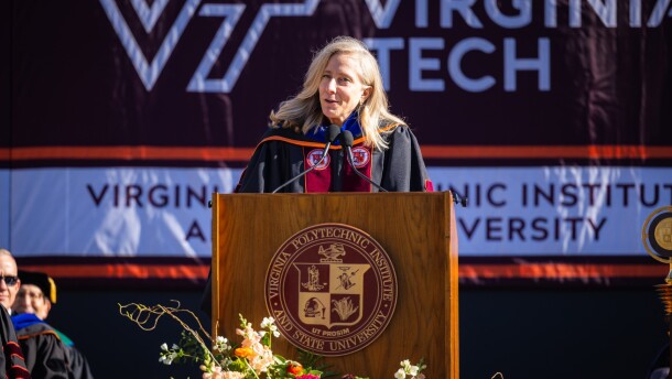 A woman with blonde hair stands at a podium, wearing a black robe with a maroon sash. 