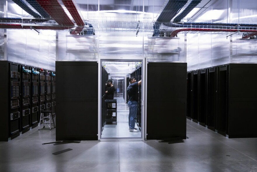 Two people work inside a glass-walled server room surrounded by tall black server racks in a bright, modern data center with overhead cabling—an environment where sustainability is prioritized through the use of hydropower to efficiently run the facility.