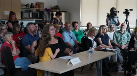 A woman sits at a table with a microphone. A seated crowd is behind her. Her daughter stands next to her.