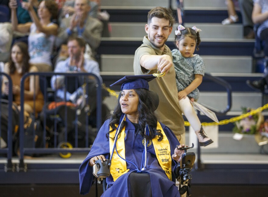 Eric Vander Kuyl turns the tassel on his wife’s, Gabrielle Vander Kuyl, mortar board with daughter Zaine Baldwin, 3, at the conclusion UNC Greensboro School of Nursing graduation Thursday, May 4, 2023. In 2016, four months before Vander Kuyl was supposed to get her Bachelor of Science in nursing, she ended up paralyzed from a tragic accident. The mother of two used online classes during the pandemic to help her finish her degree. Lynn Hey / For WUNC